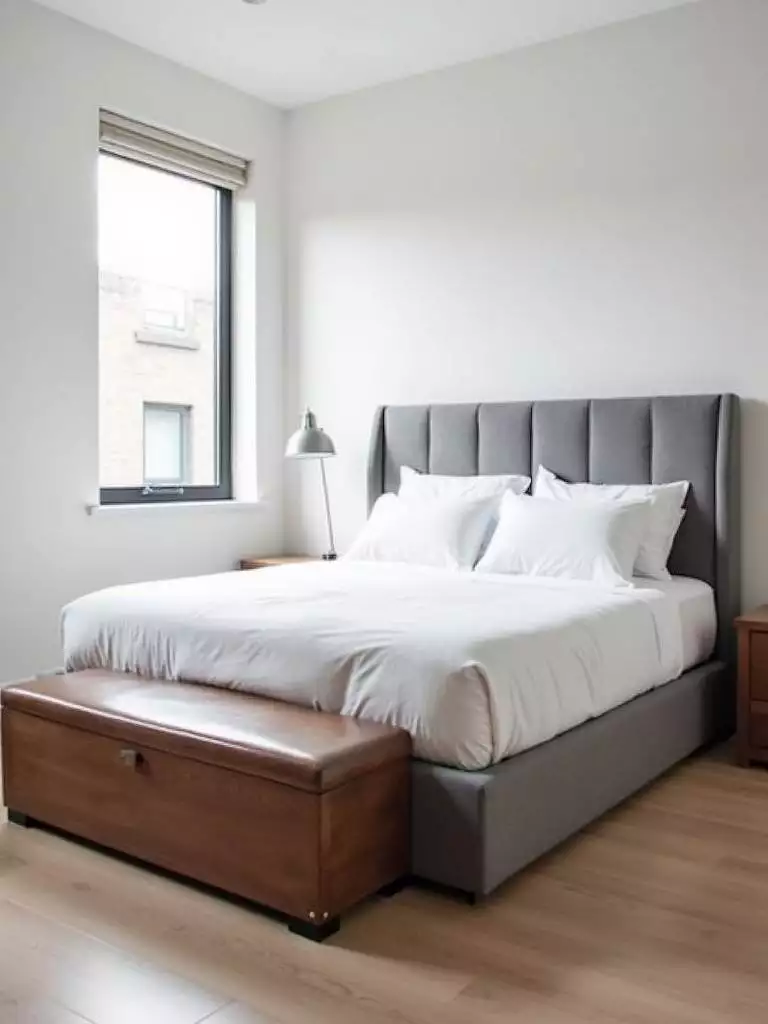 Small modern bedroom featuring a grey ottoman bed and a wooden storage bench at the foot of the bed, maximizing storage.
