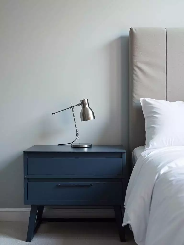 Modern bedroom bedside with a sleek dark blue nightstand, minimalist silver lamp, crisp white bedding, and soft room lighting.