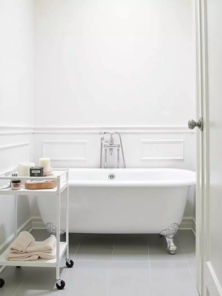 Small modern bathroom featuring a slim white metal rolling cart next to the bathtub, providing mobile and flexible storage for bath essentials in the minimalist light grey and white space.