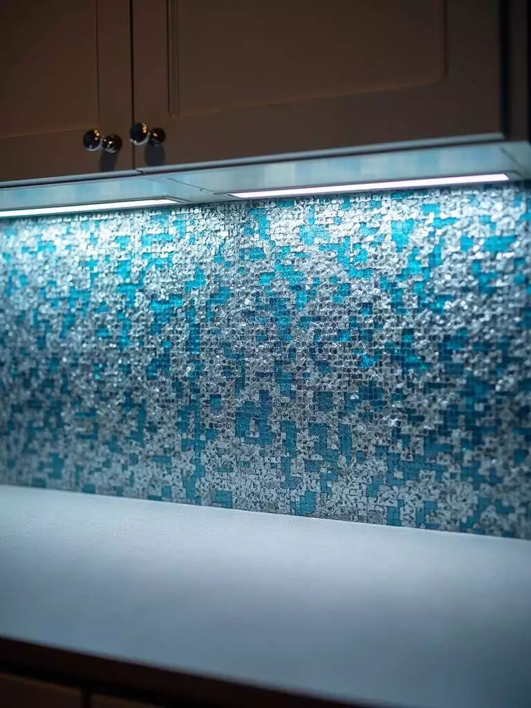 A luxurious kitchen backsplash is adorned with an intricate blue and silver glass mosaic tile pattern, softly lit by under-cabinet lighting, creating a textured and elegant focal point.