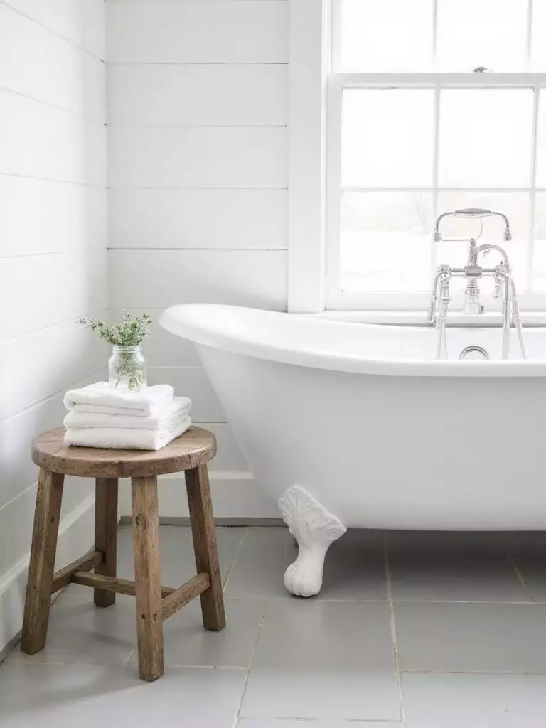 Farmhouse bathroom with white shiplap, clawfoot tub, and wooden stool holding towels.