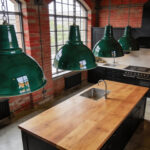 Large green vitreous enamel industrial pendant lights hanging over a wooden kitchen island in a modern British kitchen with exposed brick walls.
