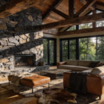 A high-end rustic living room featuring massive exposed wood beams, a large stone fireplace, and views of a forest through large windows.