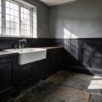 A moody and sophisticated laundry room featuring deep charcoal beadboard walls, a farmhouse sink, and slate floors inspired by historical Victorian scullery designs.