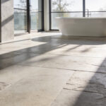A modern luxury bathroom featuring large brushed limestone floor tiles and a minimalist freestanding tub bathed in natural light.